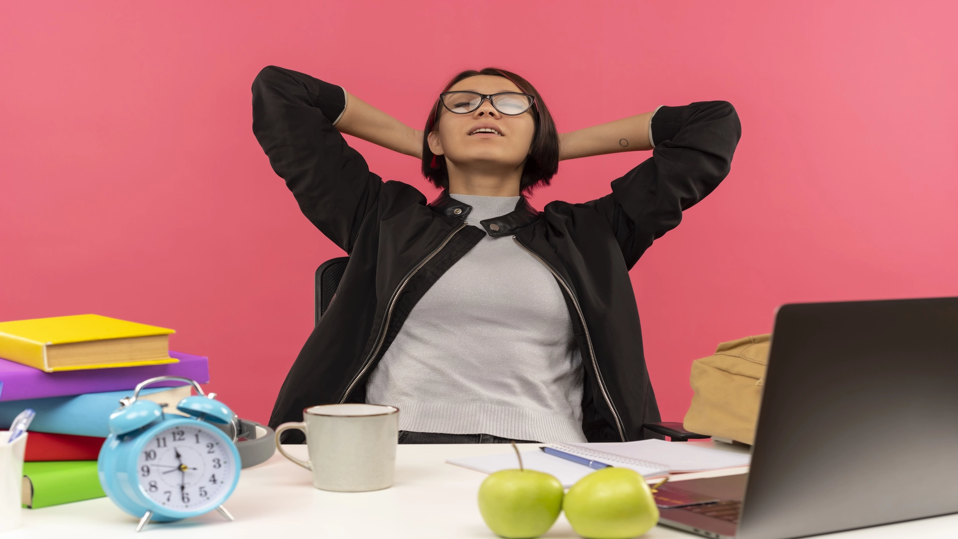 peaceful-young-student-girl-wearing-glasses-sitting-desk-putting-hands-neck-with-closed-eyes-isolated-pink-background